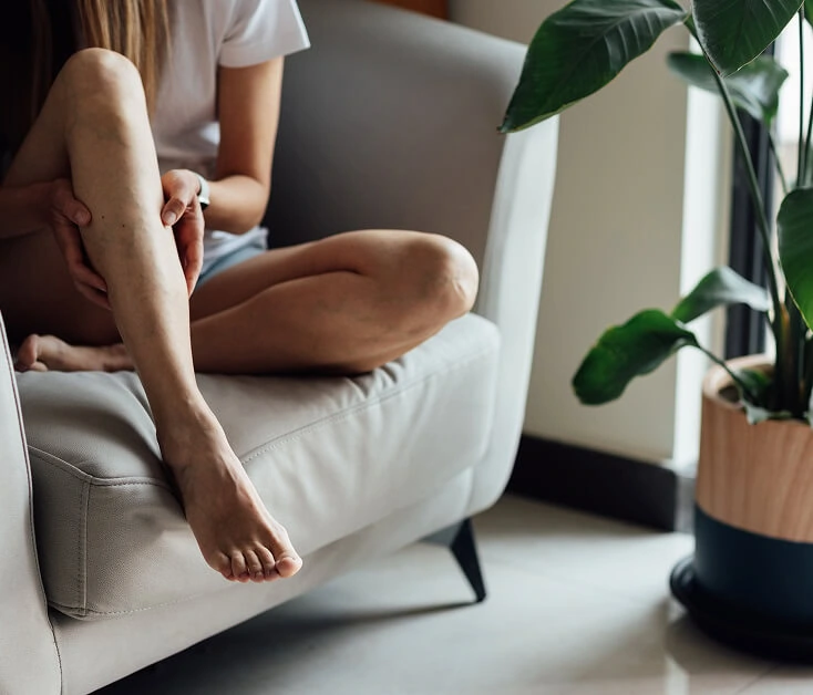 A person on a couch holds their calf, showing vein disease symptoms in Boca Raton.